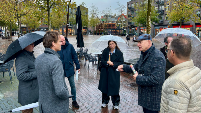 Een foto in het regenachtige centrum van stad Hardenberg. Op de foto staan 7 mensen in dichtbij in beeld. Het gaat om enkele werknemers en een wethouder van de gemeente Hardenberg, gedeputeerde Hoogland van provincie Overijssel, en een aantal van zijn werknemers.