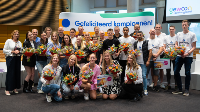 Een foto van de sporters en teams die zijn gehuldigd. Ze houden bloemen en cheques vast. Helemaal links in beeld staat wethouder Didi Dorrestijn. De groep staat in de Raadzaal van het gemeentehuis van de gemeente Hardenberg. Op de achtergrond zie je een doek met daarop de tekst "Gefeliciteerd kampioenen!"