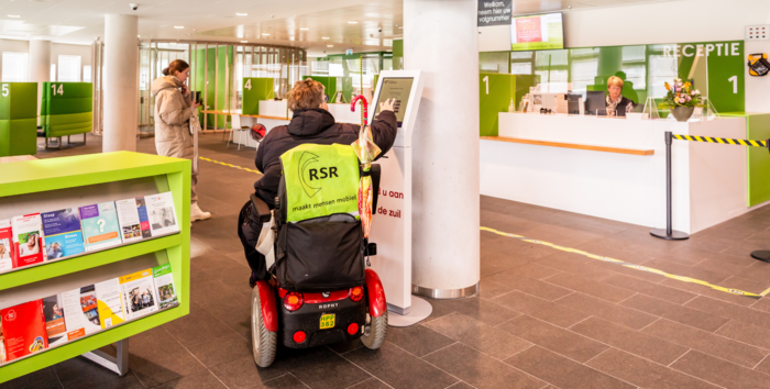 Foto van de receptieruimte in het gemeentehuis Hardenberg. U ziet een vrouw in een scootmobiel die zich aanmeldt bij de aanmeldzuil. Links van haar staat een andere vrouw te wachten in de wachtruimte. Aan de balie zit een medewerker van de gemeente Hardenberg.