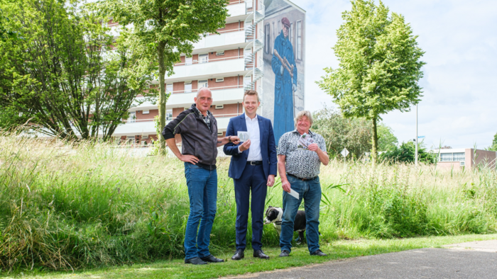 Foto van wethouder Alwin Mussche met 2 inwoners voor de muurschildering van De Klepperman (door de schilders van De Strakke Hand). De wethouder overhandigt een ansichtkaart aan de inwoner op de foto links, en de inwoner op de foto rechts houdt een aantal ansichtkaarten vast.